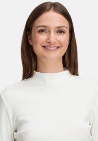 Weißes, geripptes Rollkragenoberteil mit taillierter Passform, das einen hohen Kragen und eine weiche Textur aufweist, getragen von einer Person mit langen braunen Haaren.