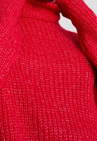 Röd stickad tröja med ett texturerat mönster. Har hög krage och långa ärmar, och visar ett mjukt, kraftigt stickat material.