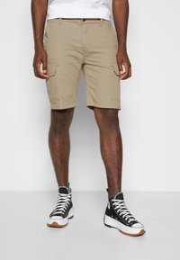 Shorts cargo beiges avec de multiples poches latérales, dotés d'une fermeture à bouton et de passants de ceinture. Associés à des baskets montantes noires.