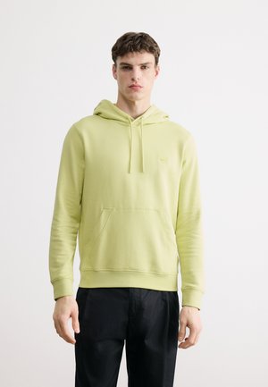 Homme portant un sweat à capuche vert clair avec poche frontale et pantalon noir, debout devant un fond clair uni.