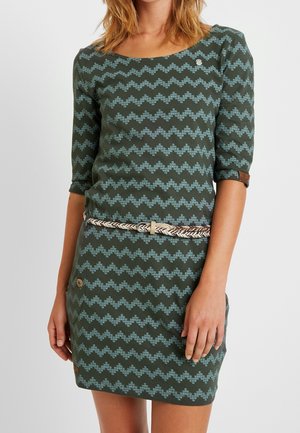 Woman wearing green dress with blue zigzag pattern, three-quarter sleeves, and braided belt at waist, standing against plain background.