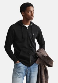 Black zip-up hoodie with drawstrings, front pockets, and logo. Model wears light blue jeans, holding a brown textured coat.