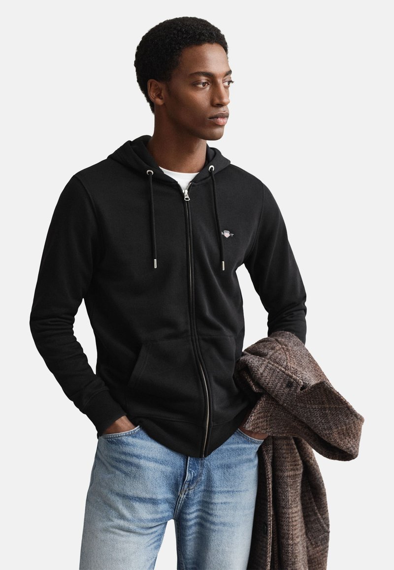 Black zip-up hoodie with drawstrings, front pockets, and logo. Model wears light blue jeans, holding a brown textured coat.