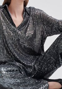 Schwarzes, paillettenbesetztes Kleidungsstück mit Kragen und Knopfleiste, locker sitzend. Reflektierende Oberfläche mit einem insgesamt schimmernden Effekt.