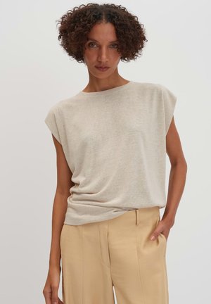 Vrouw met kort, krullend haar, gekleed in een beige mouwloze top en beige high-waist broek, staand met één hand in de zak tegen een effen achtergrond.