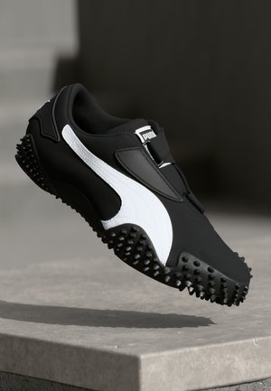 Baskets Puma noir et blanc avec semelle texturée flottant au-dessus d’un rebord en béton sur fond sombre.