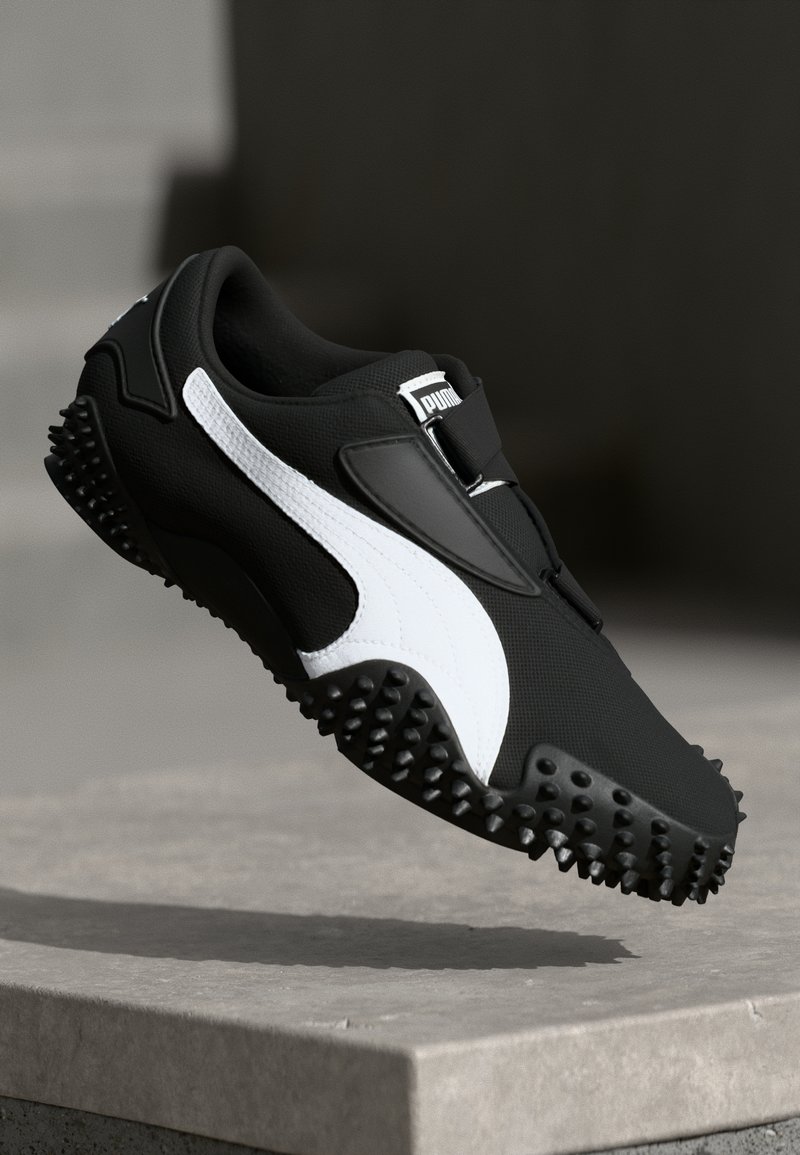 Baskets Puma noir et blanc avec semelle texturée flottant au-dessus d’un rebord en béton sur fond sombre.