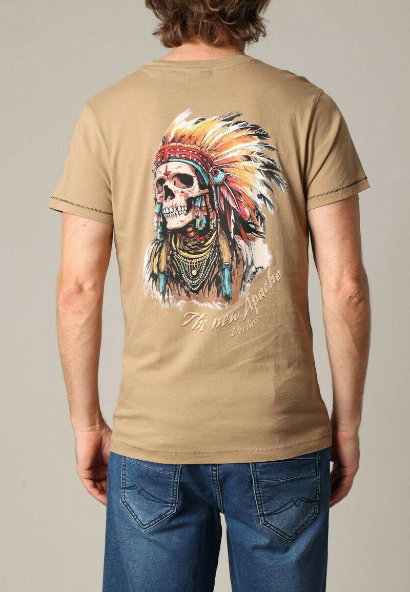 Beige katoenen t-shirt met een kleurrijke schedelafbeelding en een gevederde hoofdband, met daaronder omlijnde tekst "De nieuwe Apache."