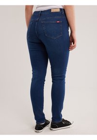 Cilvēks, kas valkā tumši zilas pieguļošas džinsa bikses un melnus sporta apavus, stāvot ar muguru pret kameru uz vienveidīga fona.