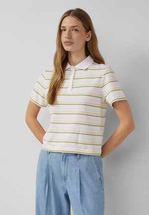 Młoda kobieta o długich jasnobrązowych włosach, ubrana w białą, krótki rękaw, koszulkę polo z zielono-żółtymi paskami i jasnoniebieskie dżinsy.