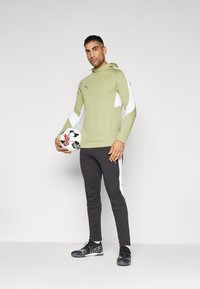 Groene hoodie met witte zijpanelen, zwarte joggingbroek en zwarte sportschoenen. Model houdt een zwart-wit voetbal vast.