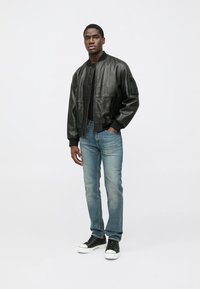 Svart läderbomberjacka med texturerad yta, ribbade manschetter och fåll, kombinerad med ljusblå jeans och svarta sneakers.