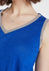 Haut bleu sans manches avec un décolleté en V, orné d'un bord argenté le long de l'encolure. Le tissu semble doux avec une finition légèrement texturée.