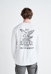 Weißes Langarm-T-Shirt mit einem schwarzen Adler-Design und dem Text "Levi's Est. 1873 California" auf der Rückseite. Aus Baumwollmaterial.