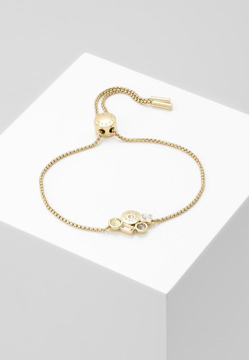 Bracelet chaîne réglable en or avec breloques en pierres précieuses rondes et carrées sur une surface d'exposition cubique blanche.