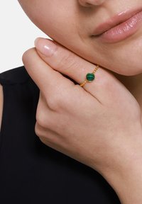 Bague en or ornée d'une pierre de malachite verte centrale avec des motifs ondulés, montée sur un anneau à billes, portée sur une main aux ongles naturels.