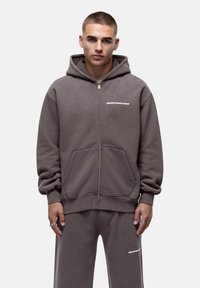Grijze zip-up hoodie van zacht materiaal, met een kangaroozak en geborduurde tekst "GOODFORNOTHING" op de linker borst.