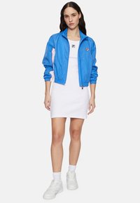 Hellblaue, leichte Zip-Jacke mit elastischen Bündchen, getragen über einem weißen gerippten Kleid. Weiße Turnschuhe und Kniestrümpfe runden das Outfit ab.