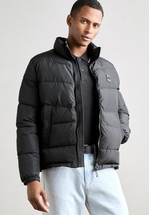 Black puffer jacket with quilted texture, high collar, zip closure, and logo patch. Worn over a black polo shirt, paired with light jeans.