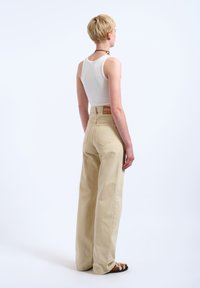 Persoon met kort blond haar, gekleed in een wit singletje, beige wijde broekspijpen en bruine sandalen, staand tegen een effen witte achtergrond.