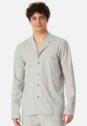 Młody mężczyzna o kręconych włosach, ubrany w jasnoszarą, pionowo prążkowaną koszulę na guziki z długimi rękawami oraz dopasowane spodnie, lekko się uśmiecha.