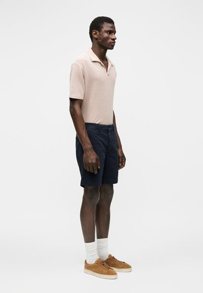 Homme debout vêtu d'un polo beige à manches courtes, d'un short bleu marine foncé, de chaussettes blanches et de baskets en daim marron sur fond blanc uni.