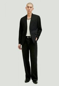 En stående person iført en sort blazer, hvid tanktop, sorte jeans, sorte sko, med en synlig brysttatovering og en neutral baggrund.