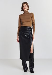 Frau in braunem Rollkragenpullover, schwarzem Lederrock mit hohem Schlitz und schwarzen kniehohen Stiefeln, die vor einem schlichten Hintergrund steht.