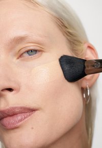 Foundation bliver påført huden med en sort børste. Ansigtet har en jævn teint, med et lys beige produkt synligt på kinden.