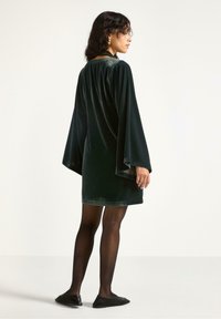 Kort kjole i mørkegrøn fløjl med brede ærmer, høj halsudskæring og en kant, der slutter midt på låret, parret med sorte strømpebukser og flade sko.