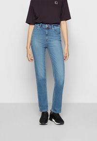 Jeans de denim de tiro alto, color azul claro, con pierna recta, diseño de cinco bolsillos y cierre de botón metálico. Combinados con zapatillas deportivas negras.