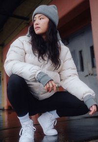 Light beige puffer jacket with a zip, gray cuffs, black leggings, and white sneakers. The background features an industrial setting.