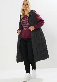 Svart vadderad lång väst med huva, med dragkedja framtill och sidofickor, bärs över en burgundy sweatshirt och svarta byxor.