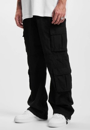 Homme portant un pantalon cargo noir à multiples poches et des baskets blanches, debout les mains détendues le long du corps.