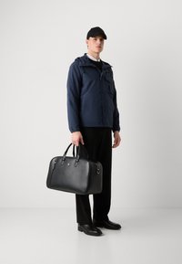 Jachetă impermeabilă de culoare bleumarin, cu glugă și buzunare; geantă de voiaj din piele neagră, cu mânere duble; pantaloni și cizme negre; fundal simplu.