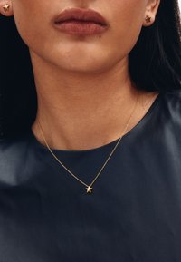 Goldene Stern-Halskette mit einer filigranen Kette, getragen über einem dunkelblauen, strukturierten Kleidungsstück, ergänzt durch kleine goldene Stern-Ohrringe.