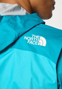 Artimu matomas žmogaus petys, vilkintis ryškiai mėlyną striukę su dideliu baltu „The North Face“ logotipu.