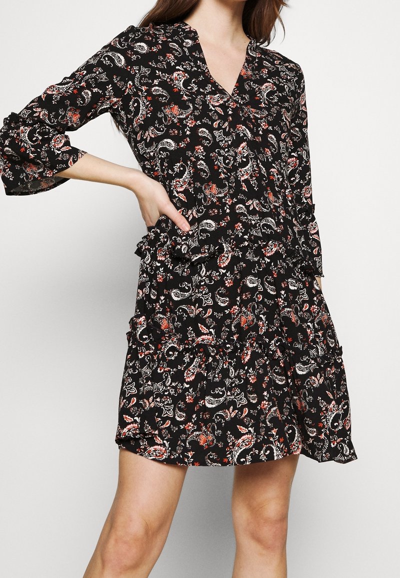 Femme portant une robe noire à motif floral avec des manches longues et un col en V, debout, une main sur la hanche, devant un fond uni.