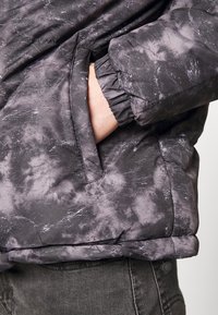 Hand som är stoppad i sidofickan på en grå och svart marmorering puffjacka med elastisk mudd, bärs över mörka jeans.