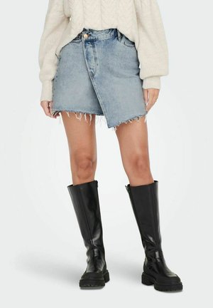 La gonna mini in denim presenta un colore azzurro chiaro sbiadito, orlo sfrangiato e un design asimmetrico avvolgente. Abbinata a stivali neri di pelle alti.