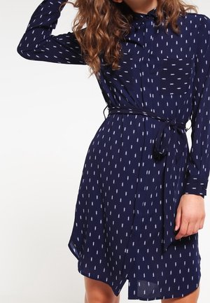 Sukienka koszulowa w kolorze granatowym z białym geometrycznym wzorem, długimi rękawami, kołnierzykiem i paskiem w talii. Lekka tkanina o zwiewnym kroju.