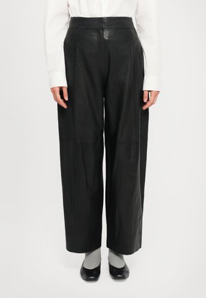 Pantaloni in pelle nera a vita alta con gamba ampia, texture liscia e cuciture a contrasto, abbinati a calzini grigi e scarpe nere.