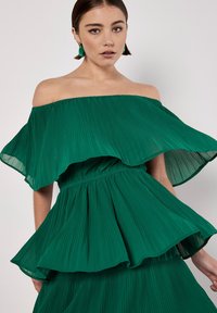 Grünes plissee Kleid mit schulterfreiem Design, geschichteten Rüschen und strukturiertem Stoff. Verfügt über eine taillierte Taille und eine fließende Silhouette.
