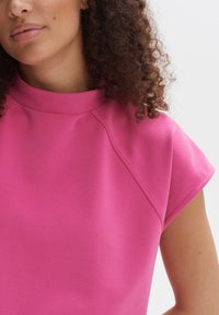 Roze top met korte mouwen en een hoge hals, met een gladde textuur en raglanmouwen, die een gestructureerd en modern ontwerp benadrukt.