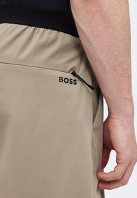 Κοντινό πλάνο μπεζ σορτσάκι με μαύρη ελαστική μέση και φερμουάρ τσέπη με την ετικέτα "BOSS" κοντά στο χέρι ενός ατόμου.