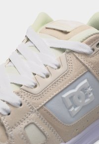 Αθλητικό παπούτσι DC σε μπεζ και άσπρο με άσπρα κορδόνια, ραμμένα πάνελ από σουέτ, πλέγμα στις πλευρές και ανάγλυφο λογότυπο DC στην εξωτερική πλευρά.