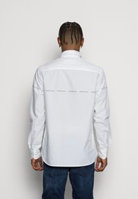 Vit långärmad skjorta med knäppt krage. Har "Calvin Klein Jeans"-logotypen på baksidan och en rak fåll. Slät bomullstextur.