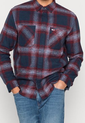 Man wearing blue, red, and white plaid long-sleeve button-up shirt with two chest pockets and blue jeans, hands in pockets.