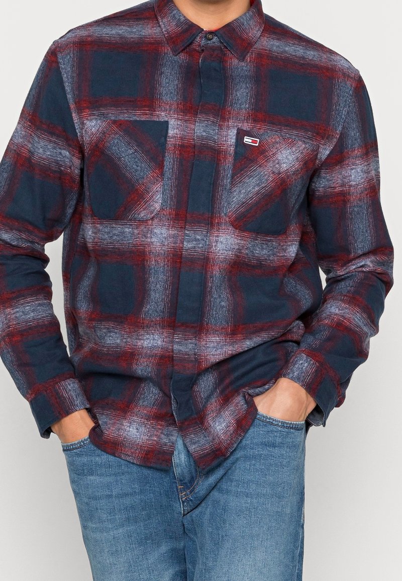 Homme portant une chemise à carreaux bleue, rouge et blanche à manches longues avec deux poches poitrine et un jean bleu, les mains dans les poches.