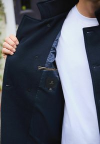 Marineblauer Mantel mit strukturierter Außenseite, ausgestattet mit einem glatten Satin-Innenfutter, Knopfdetails und einer Brusttasche mit Etikett.